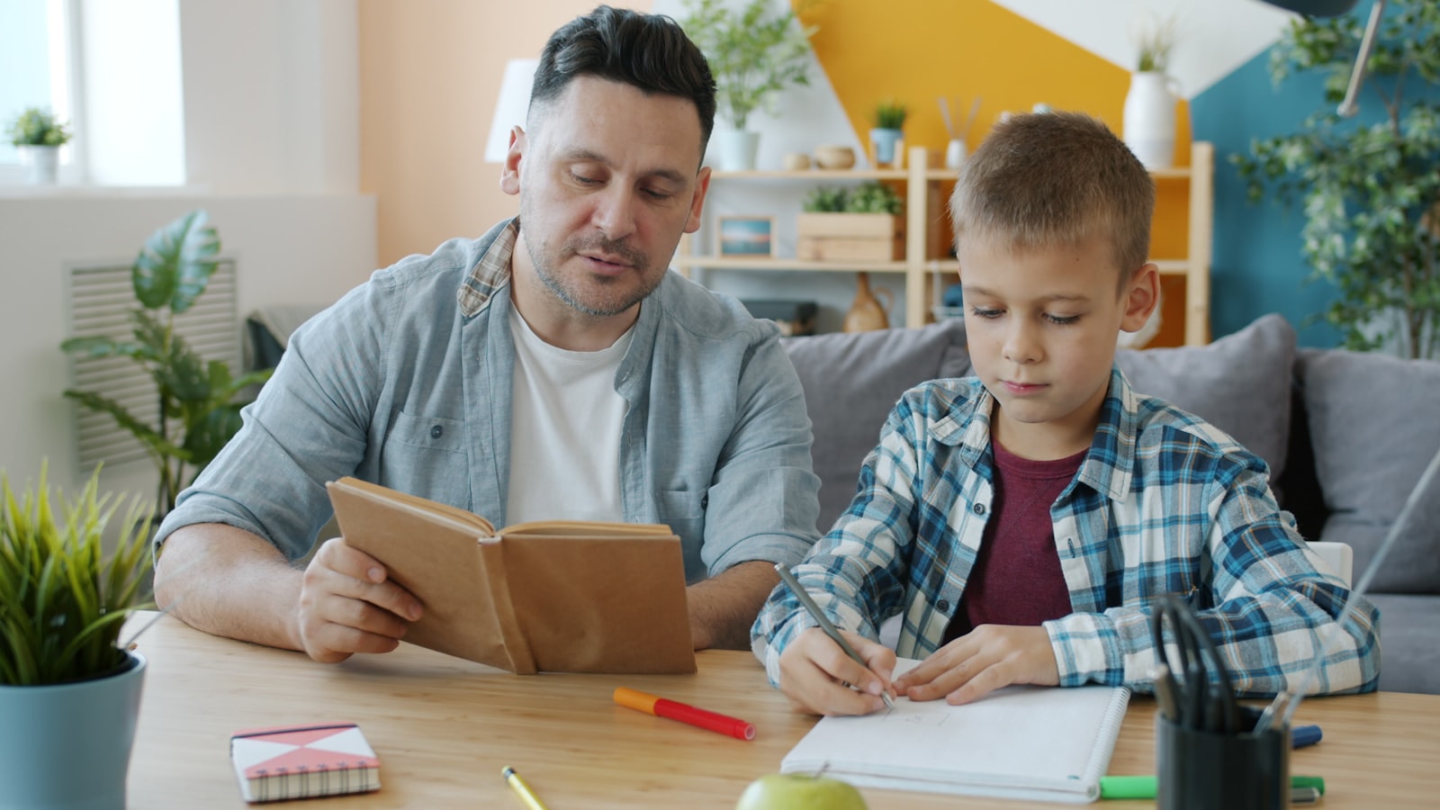 A father helping his young child fill out a school registration form at the kitchen table